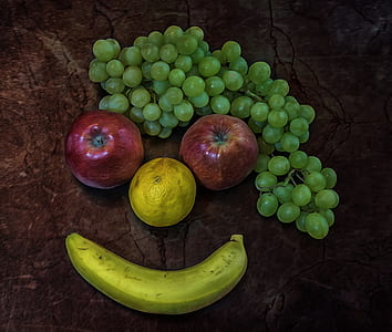 red apples, round yellow fruit, green grapes, and green banana forming face on brown surface thumbnail