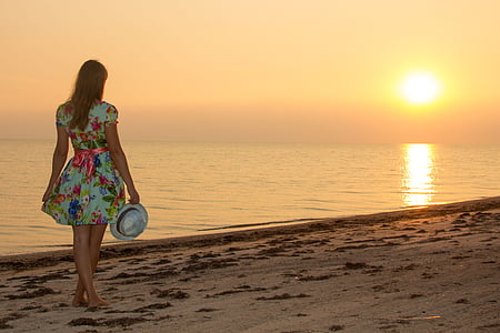 woman in teal, blue, and red floral dress standing on seashore while holding white fedora hat thumbnail