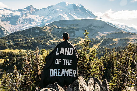 man wearing black and of the dreamer printed cape while standing on gray mountain overlooking forest with green trees at daytime thumbnail