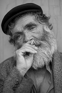 grayscale photo of old man with beard wearing hat holding cigarette stick thumbnail