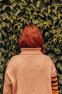 woman with red hair in gray, orange, and black sweater facing on green plant thumbnail