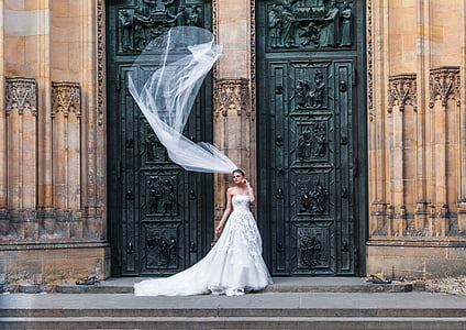woman wearing wedding dress standing in front of brown and black building while wedding veil's floating on midair thumbnail
