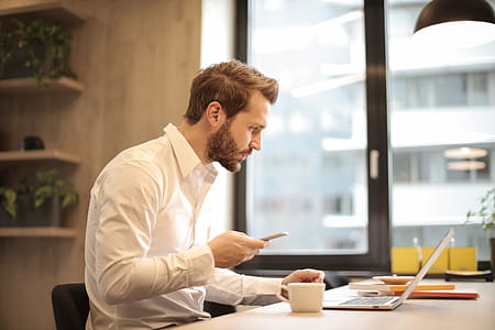 A young bearded caucasian man in formals looking at his laptop screen while holding his mobile phone thumbnail
