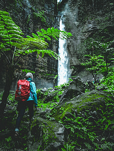 person wearing blue windbreaker jacket and black pants carrying red backpack near waterfalls thumbnail