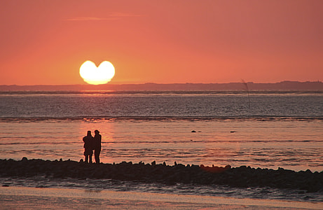 silhouette photo of couple standing near shore facing heart sun thumbnail