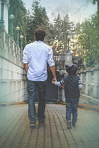 man in white collared long-sleeved shirt holding boy in black leather hooded jacket walking near green leaf trees during daytime thumbnail