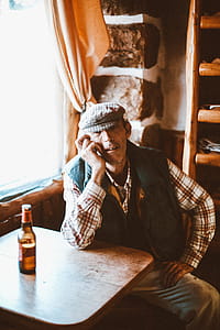 Man Wearing Red and White Plaid Shirt and Black Vest Sitting Beside Brown Wooden Table thumbnail