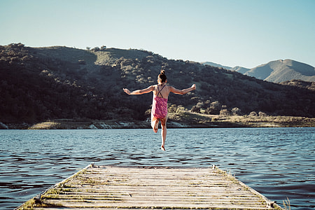 woman jumping from a wooding dock towards a lake surrounded by mountains during daytime thumbnail