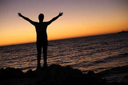 silhouette photo of person beside seashore raising hand during sunset thumbnail