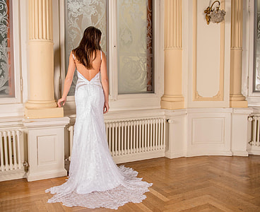 woman in white backless wedding gown standing beside a beige concrete pillar inside a room with floral-etched glass window panels thumbnail