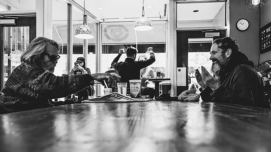 grayscale photo of two men sitting beside table inside building thumbnail