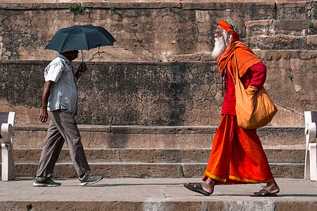 man in orange and red dress walking towards man holding umbrella during daytime thumbnail