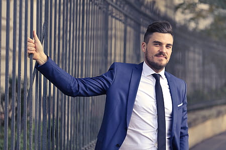 A bearded caucasian man in blue formal suit and white shirt standing against metal railings thumbnail