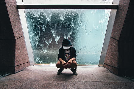person wearing hoodie and fitted cap squatting in front of glass wall with water flowing photo thumbnail