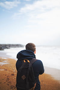 man wearing backpack facing body of water under white sky during daytime thumbnail