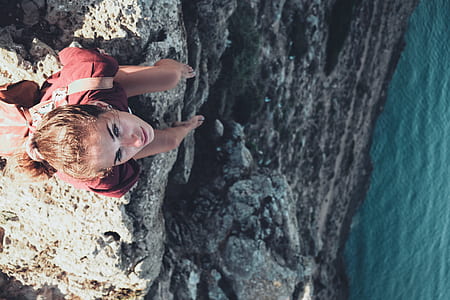 aerial photo of woman sitting on cliff near body of water during daytime thumbnail