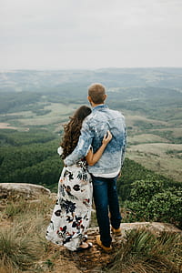 man in blue denim jacket beside woman in black and white floral dress standing in front of mountains during daytime thumbnail
