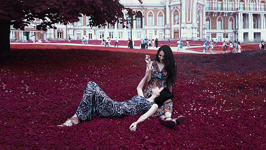 woman in black floral dress sleeping on woman's lap in brown floral dress while sitting on red grass thumbnail