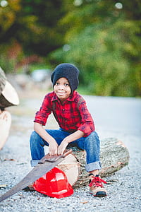 boy wearing red and black plaid dress shirt sitting on wood trunk while holding hand saw outdoors thumbnail