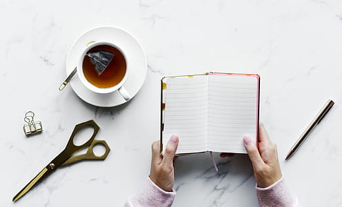 Person Holding Empty Book Near Pen and Shears With a Cup of Tea thumbnail