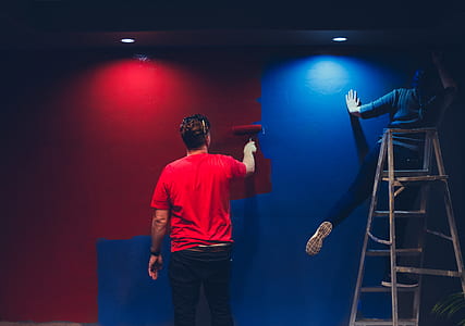 man standing on ladder while painting a blue paint on wall beside man painting a red on the same wall thumbnail