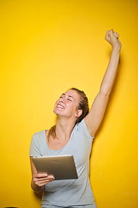 Excited Young Woman Holding An iPad With One Hand Up Against The Yellow Wall thumbnail