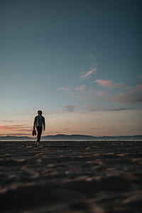 man walking barefoot on sand covered field while carrying shoes during day thumbnail