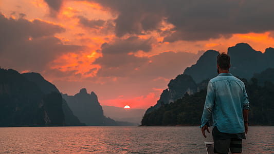 man wearing blue chambray shirt facing body of water with view of mountains during sunset photo thumbnail