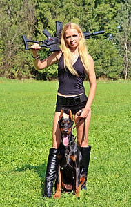 woman wearing black tank top with black short shorts while holding black assault rifle and black and rust Pinscher Doberman thumbnail