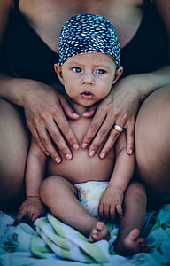 woman behind baby wearing diaper and blue hat sitting on white and green textile thumbnail