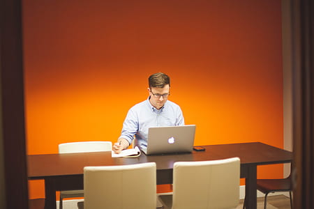 man in blue dress shirt wearing eyeglasses sitting on chair while looking at the silver iMac and holding pen thumbnail