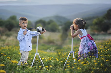 selective focus photography of boy and girl on flower field using while standing near cameras with tripods thumbnail