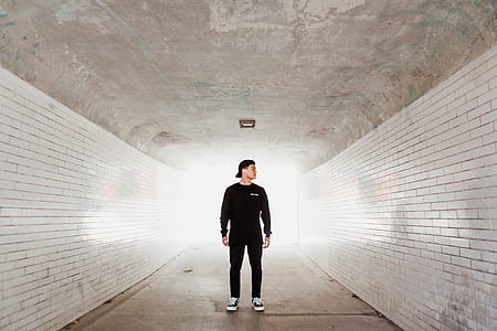 man standing at the center of pathway between white concrete brick wall bricks looking at left side during daytime thumbnail