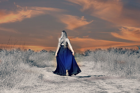 woman in navy-blue cage-back floor-length dress walking through the rocky road surrounded with grass during golden hour thumbnail