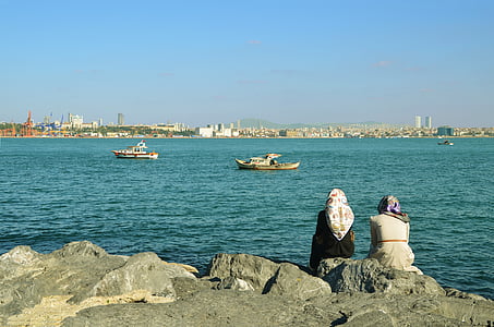 two women sitting on stone beside water thumbnail
