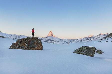 man wearing red jacket and black pants standing on brown rock in the middle of snow covered field at daytime thumbnail