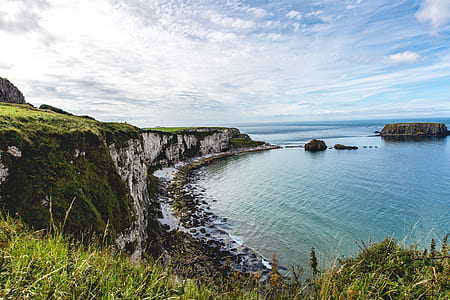 seashore near mountain cliff covered in green grass overlooking islet under blue sky at daytime thumbnail