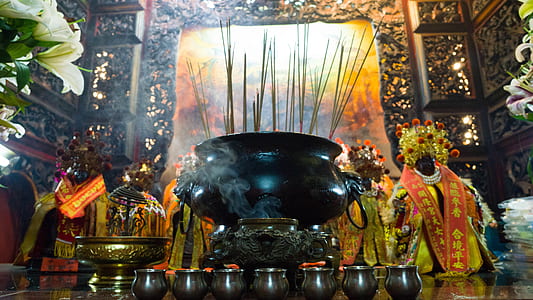 long-angle photo of incense sticks struck on a black cauldron in front of a painting in a well-lit temple thumbnail