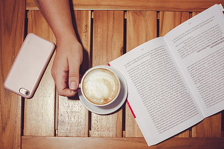Person's Hand Holding White Coffee Mug With Plate on Brown Wooden Board With White and Black Book thumbnail