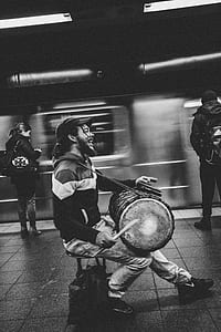 greyscale photography of man playing drum on train station beside two person waiting for train thumbnail