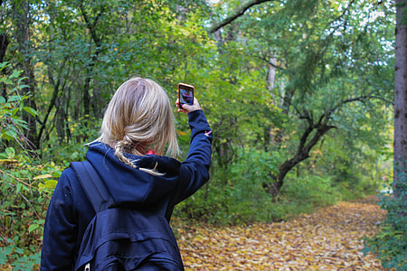 woman blonde haired wearing blue hoodie and black backpack while taking selfie in forest during daytime thumbnail