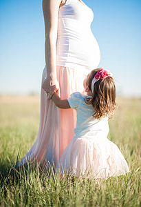 girl in white and pink puff sleeve dress near pregnant mother standing on green grass during daytime thumbnail