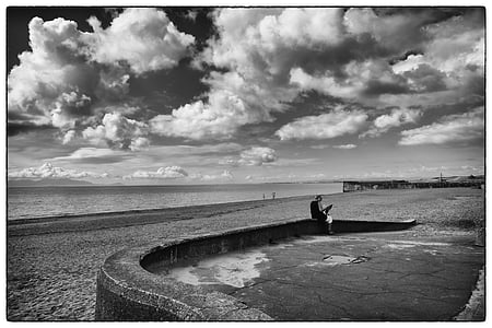 grayscale photography of person sitting on concrete barrier near seashore thumbnail