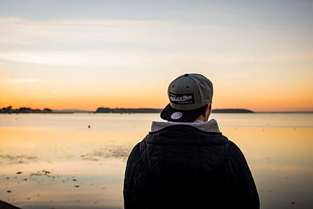 man wearing black jacket and gray flat-brim cap standing near body of water thumbnail