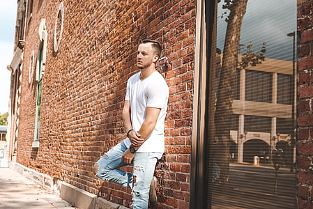 man in white crew-neck shirt and blue denim pants leaning on brown concrete wall during daytime thumbnail