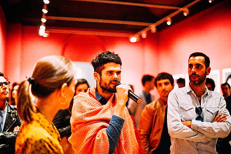 man in blue top holding black microphone surrounded with crowd inside red painted room thumbnail