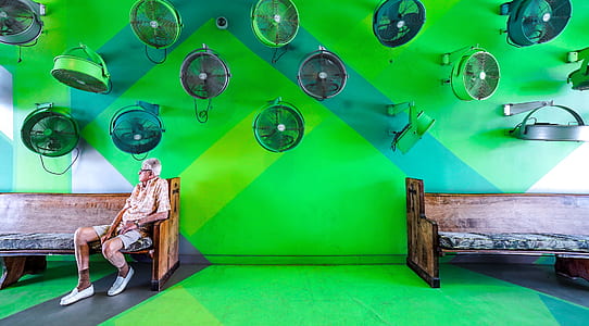 man sitting on brown wooden bench inside green concrete room with gray and black wall fans thumbnail