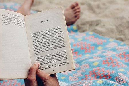 person holding book on white sand with blue blanket during daytime thumbnail