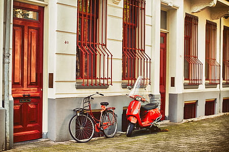red city bike leaning on white concrete building beside red motor scooter during daytime thumbnail