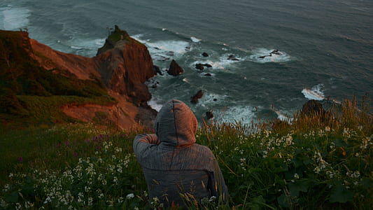 person in hoodie seating on field of flower in front of body of water thumbnail